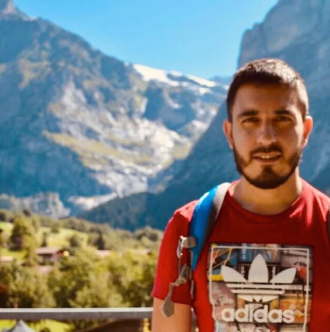 Felipe in a red T-shirt with a backpack strap, in front of snow-capped mountains and a green valley