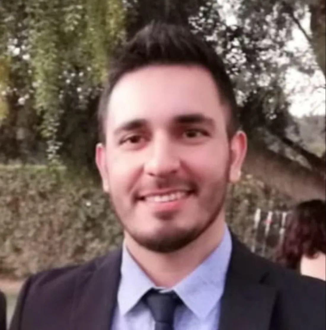 Felipe smiling outdoors, wearing a dark suit, light blue shirt and tie